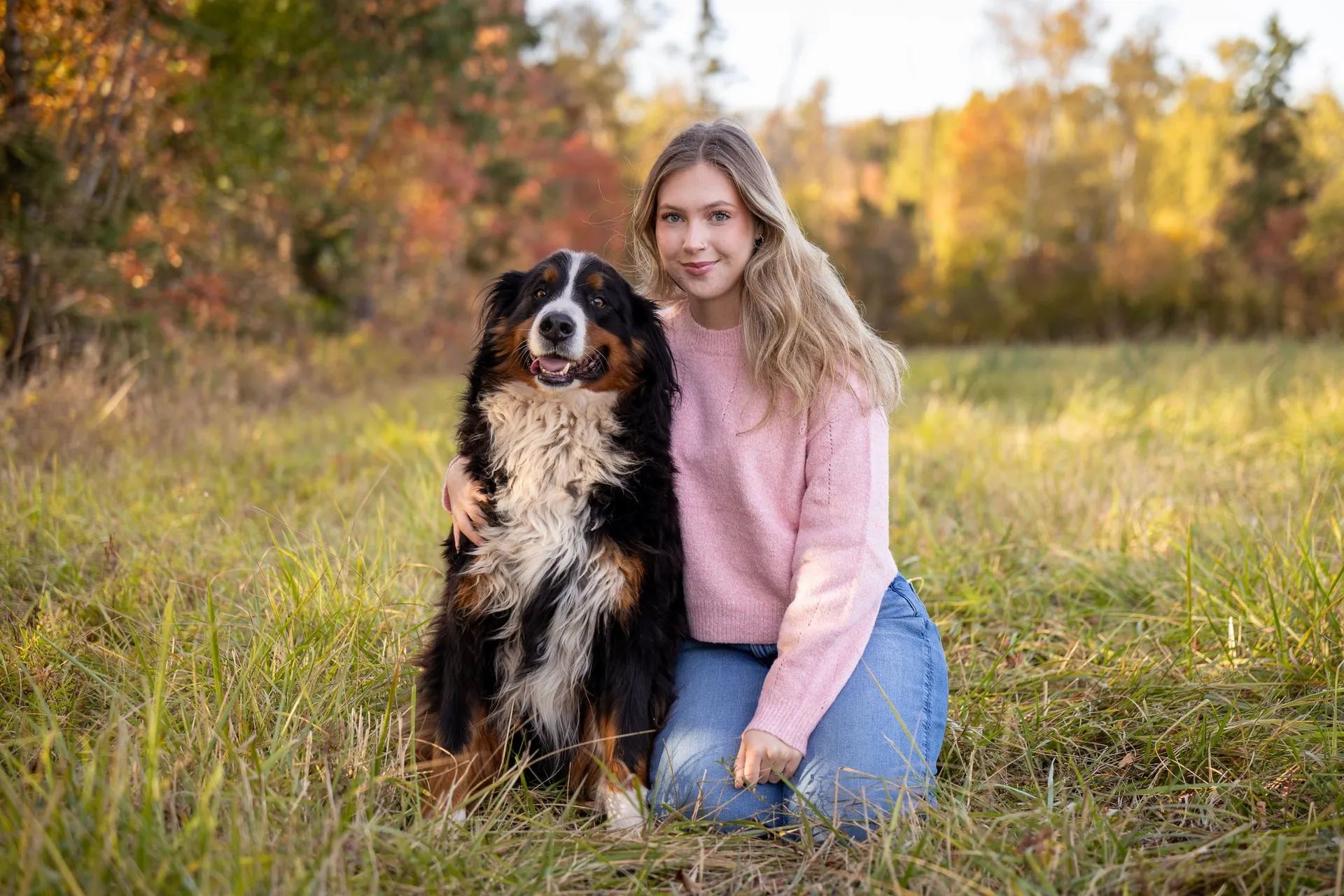 A young woman kneeling in an autumn field beside her Bernese Mountain Dog